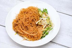 Vermicelli di noodles fritti su piatto bianco, noodle fritti in stile cinese con germogli di soia e coriandolo su tavola di legno - cibo tailandese foto