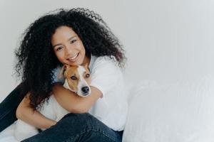 foto di una giovane donna attraente con un taglio afro, abbraccia con amore il cane, si prende cura degli animali domestici, sorride dolcemente, indossa abiti casual, isolata su sfondo bianco, si siede sul letto, copia lo spazio per il tuo promo