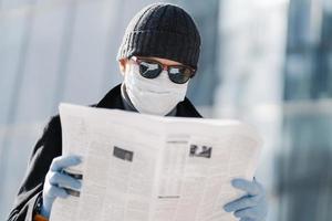 la foto dell'uomo europeo indossa una maschera medica protettiva e guanti, cammina per la città all'aperto, legge un articolo sull'epidemia di virus in diversi paesi utilizza misure di protezione durante la diffusione del coronavirus