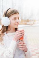 giovane ragazza che beve caffè in un parco. adolescente che indossa le cuffie e tiene il caffè per andare. portare via il concetto di bevanda. vista laterale foto