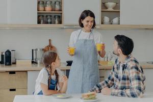 l'adorabile madre felice indossa il grembiule, tiene due bicchieri di succo d'arancia, parla con marito e figlia, prepara la colazione. tre membri della famiglia cenano in cucina. concetto di stare insieme. foto