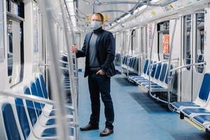 scatto a figura intera di imprenditore maschio in abito formale, maschera medica, sta in treno urbano durante l'epidemia di malattia, protezione dai virus e quarantena si protegge dal coronavirus sui trasporti pubblici foto