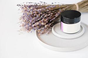 Crema viso alla lavanda in un vasetto senza marchio su vassoi di cemento con fiori di lavanda sullo sfondo. copia spazio. ingrediente calmante e lenitivo per aromaterapia e cure di bellezza foto