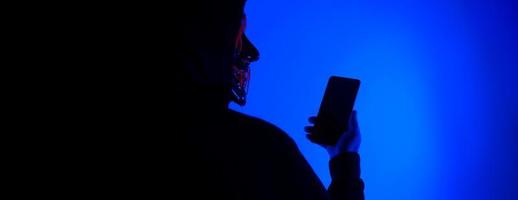 hacker anonimo e maschera facciale con smartphone in mano. foto