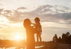 una silhouette di una bambina felice le braccia della sua amorevole madre per un abbraccio, davanti al tramonto nel cielo in una giornata estiva. foto