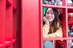 bella donna asiatica dai capelli lunghi che indossa una veste gialla e un sorriso felice in piedi nella cabina telefonica rossa nel tema della celebrazione del natale e del felice anno nuovo foto