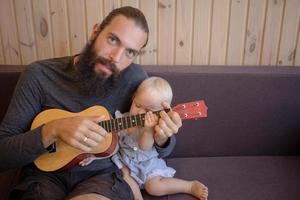 il padre barbuto con i bambini gioca sull'ukulele al coperto foto