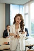Ritratto di giovane donna asiatica d'affari in piedi in ufficio con le braccia incrociate con un collega di colleghi che parla in background foto