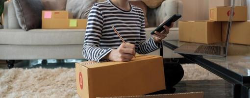 giovane donna piccolo imprenditore shopping online a casa. confermare gli ordini dei clienti con telefoni cellulari che preparano il pacchetto di prodotti in background. piccolo imprenditore o concetto di stile di vita freelance. foto