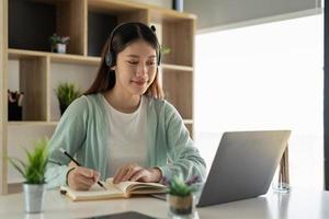 giovane donna asiatica che scrive facendo la lista che prende appunti nel blocco note che lavora o impara sul computer portatile all'interno - corso educativo o formazione, seminario, concetto online di istruzione foto