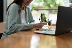 giovane studentessa asiatica indossa le cuffie che scrivono sul taccuino per studiare la lingua online guarda e ascolta il docente, webinar tramite videochiamata e-learning a casa foto