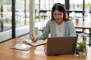 donna asiatica che scrive lista che prende appunti nel blocco note lavorando o imparando su laptop al chiuso - corso educativo o formazione, seminario, concetto online di istruzione foto