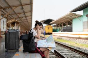 giovane donna viaggiatore con zaino che cerca di mappare in attesa del treno, viaggiatore con zaino e sacco a pelo asiatico sulla piattaforma ferroviaria alla stazione ferroviaria. concetto di vacanza, viaggio, viaggio e viaggio estivo estivo foto