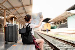 giovane donna viaggiatore con zaino che cerca di mappare in attesa del treno, viaggiatore con zaino e sacco a pelo asiatico sulla piattaforma ferroviaria alla stazione ferroviaria. concetto di vacanza, viaggio, viaggio e viaggio estivo estivo foto