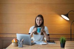 vista frontale sullo studio della giovane donna asiatica davanti al computer portatile a casa - libro di lettura della ragazza che prepara il test d'esame con consultazione online - concetto di istruzione e apprendimento. foto