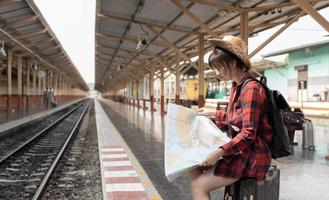 giovane donna viaggiatore con zaino che cerca di mappare in attesa del treno, viaggiatore con zaino e sacco a pelo asiatico sulla piattaforma ferroviaria alla stazione ferroviaria. concetto di vacanza, viaggio, viaggio e viaggio estivo estivo foto