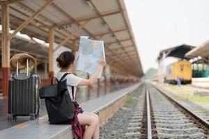 giovane donna asiatica che utilizza una mappa locale generica, seduta da sola al binario della stazione ferroviaria con i bagagli. vacanze estive in viaggio o concetto di giovane viaggiatore zaino turistico foto