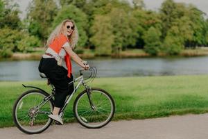 scatto all'aperto di felice donna bionda riccia indossa occhiali da sole, vestito con abiti casual attivi, va in bicicletta vicino al lago e alberi verdi in campagna, trascorre il tempo libero all'aperto, ama l'hobby preferito foto