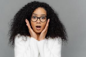 omg, non può essere così sorpresa emotiva giovane femmina dalla pelle scura con i capelli ricci, tocca entrambe le guance, ha un'espressione facciale scioccata, vestita con un maglione bianco, isolata su sfondo grigio foto