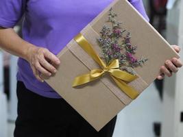 donna con scatola regalo di colore marrone avvolto in carta fiore viola secco con ciotola di nastro d'oro, regali di festival per natale e felice anno nuovo foto