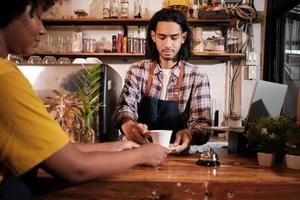 di fronte al primo piano, un giovane barista maschio tailandese dai capelli lunghi lavora servendo una tazza di caffè a una cliente afroamericana al bancone della caffetteria, un buon lavoro di servizio e una piccola imprenditrice. foto