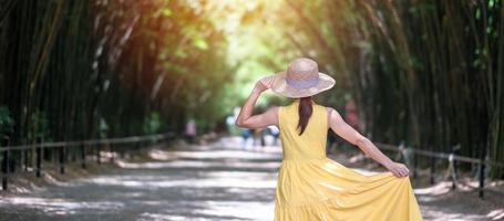 donna asiatica in abito giallo e cappello che viaggia nel tunnel di bambù verde, viaggiatore felice che cammina nel tempio di chulabhorn wanaram. punto di riferimento e popolare per le attrazioni turistiche a nakhon nayok, in tailandia foto