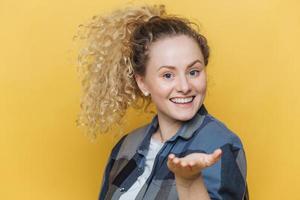 bella donna felice allunga la mano in primo piano, ha un ampio sorriso, mostra denti bianchi perfetti, indossa una camicia a scacchi, posa su sfondo giallo. sorridente soddisfatta modello femminile indoor foto