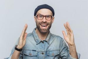 bell'uomo felice alza le mani con eccitazione, ha un'espressione felicissima, guarda con una faccia felice, ha folta barba e baffi. uomo felice in occhiali pone su sfondo bianco, si diverte foto