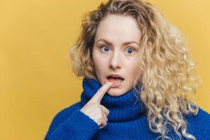 colpo isolato di bella donna stupita con capelli biondi ricci, tiene il dito anteriore sul labbro inferiore, ascolta attentamente qualcuno, vestito con un maglione blu, isolato su sfondo giallo. emozioni foto