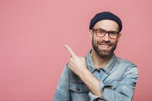 colpo orizzontale di allegro uomo barbuto con espressione felice, vestito con abiti alla moda, indossa occhiali, cappello e camicia di jeans, indicato con il dito anteriore nello spazio vuoto della copia per la tua pubblicità foto