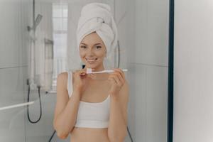 felice bella signora con un sano sorriso perfetto lavarsi i denti mentre si è in piedi in bagno foto