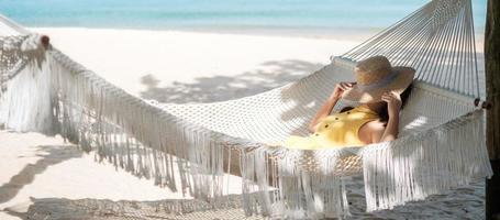 il viaggiatore felice della donna si sta rilassando su un'amaca sulla spiaggia paradisiaca. turista femminile in abito giallo riposa vicino al mare tropicale. concetto di vacanza, viaggio, estate, voglia di viaggiare e vacanza foto