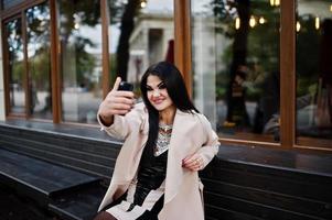 donna sexy capelli neri in occhiali e cappotto seduto su una panchina con il telefono cellulare a portata di mano e fare selfie. foto