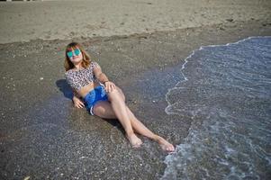 bellissima modella che si rilassa su una spiaggia di mare, indossa jeans corti, camicia leopardata e occhiali da sole. foto