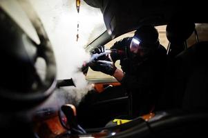 uomo in uniforme e respiratore, lavoratore del centro di autolavaggio, pulizia degli interni dell'auto con pulitore a vapore caldo. concetto di dettagli dell'auto. foto