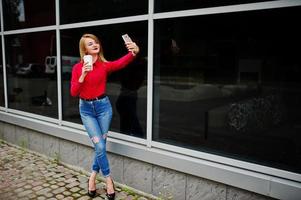 ritratto di una bella donna in camicetta rossa e jeans casual che si fa selfie sul cellulare e tiene una tazza di caffè fuori dall'enorme centro commerciale. foto