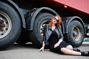 ragazza elegante dai capelli rossi indossa in nero, seduta contro grandi ruote di camion. foto