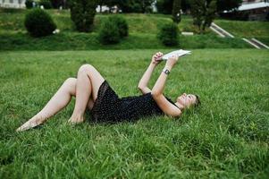 ritratto di una bella donna in abito nero a pois sdraiato sull'erba e leggendo un libro nel parco. foto