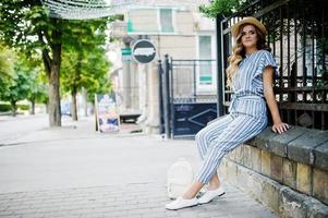 ritratto di una bella donna in tuta a righe e cappello seduto e in posa su una recinzione in una strada della città. foto