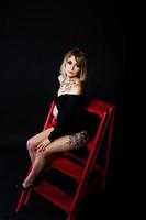 ritratto in studio di ragazza bionda con trucco originale sul collo e tatuaggio sulla coscia, indossa un abito nero su sfondo scuro, seduto su una scala rossa. foto