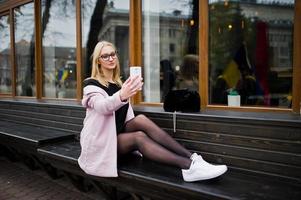 ragazza bionda con occhiali e cappotto rosa, tunica nera seduta su una panchina in strada e facendo selfie dallo smartphone. foto