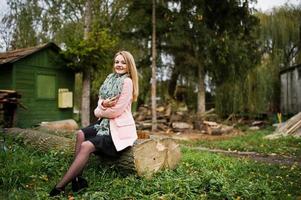 giovane ragazza bionda al cappotto rosa posata sul parco autunnale, seduta su un albero tagliato contro la casa del guardaboschi. foto