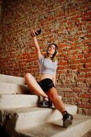 la ragazza indossa pantaloncini con grandi cuffie per fare selfie in una fabbrica abbandonata con muri di mattoni. foto