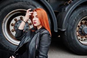 ragazza elegante dai capelli rossi indossa in nero, seduta contro grandi ruote di camion. foto