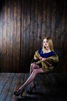 elegante ragazza bionda indossare sul cappotto di pelliccia seduto su una sedia in studio su sfondo di legno. foto