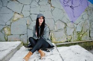 elegante ragazza bruna con berretto grigio, casual street style con skate board in una giornata invernale contro la parete colorata. foto