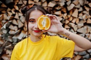 giovane ragazza divertente con trucco luminoso, indossare su camicia gialla tenere un pezzo di arancione su sfondo di legno. foto