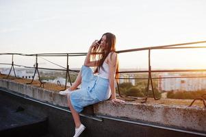 ritratto di una splendida giovane donna in maglietta bianca e gonna blu seduta sul bordo dell'edificio al tramonto. foto