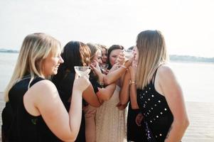 un gruppo di 8 ragazze indossano il nero e 2 spose alla festa di addio al nubilato contro la spiaggia soleggiata bevendo champagne. foto