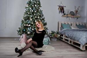 ragazza bionda carina in abito nero contro l'albero di Capodanno con regali di Natale in studio. Buone vacanze. foto